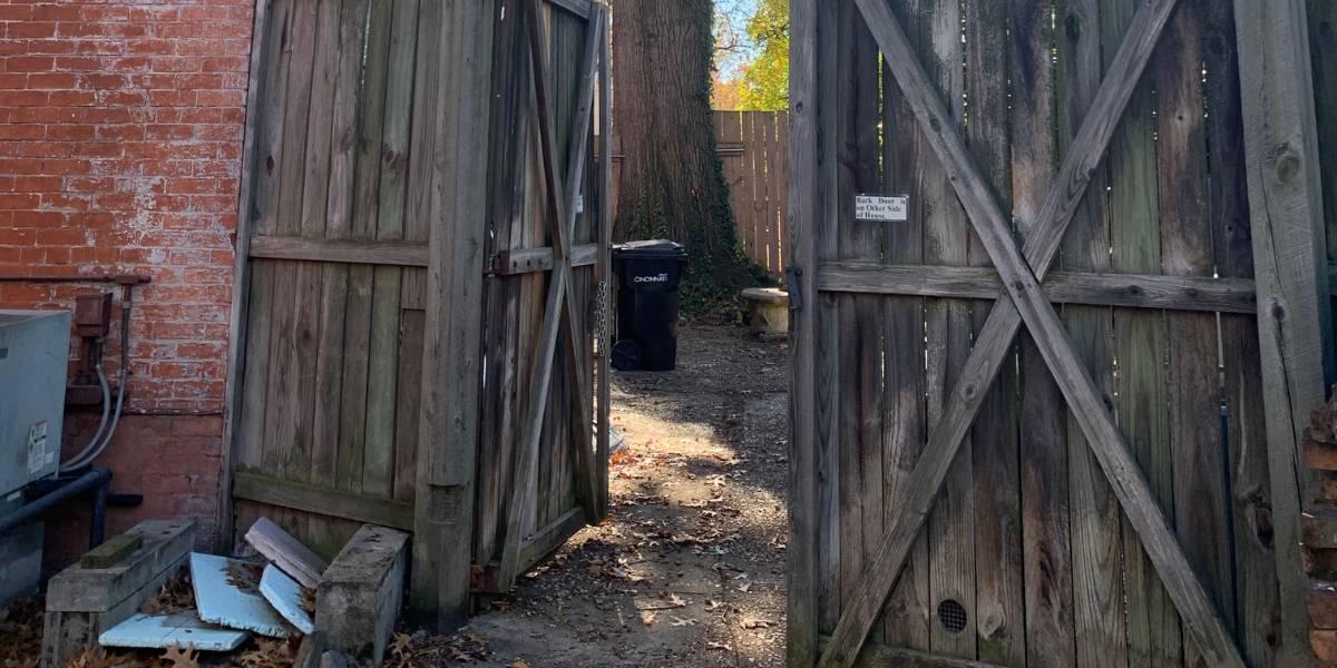 An open wooden gate shows a difficult to access yard with a large tree that needs to be removed growing very close to a red brick home. This displays the difficulty of a confined space tree removal.